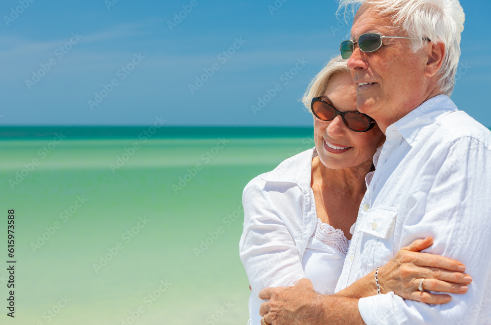 Happy senior man and woman retired couple embracing wearing sunglasses on a deserted tropical beach with turquoise sea and clear blue sky