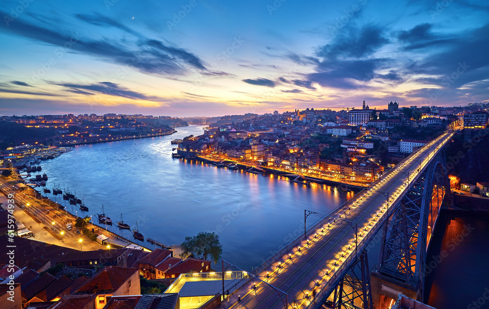 Porto, Portugal. Evening sunset panoramic view at nighttime town and Ponte de Dom Luis bridge with tramways. Coastline of river Douro with reflections of illumination in water and picturesque clouds o