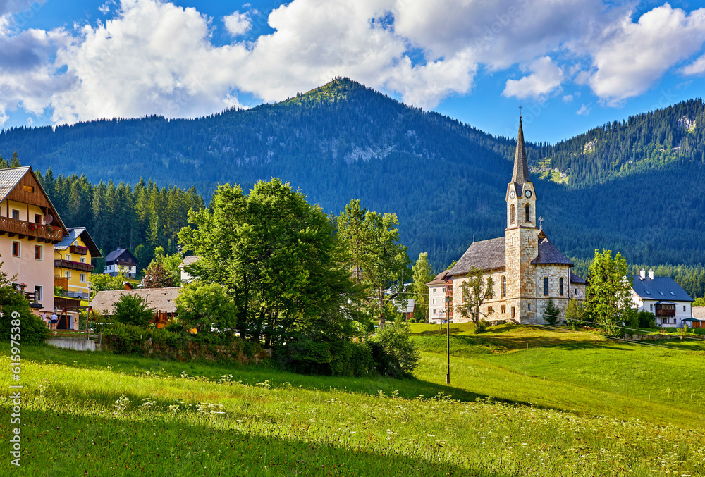 Austria. Traditional church with chapel in village among picturesque landscapes in austrian Alps mountains. Summer green lawns and fields. Knolls covered with forests and trees. Blue sky with clouds.