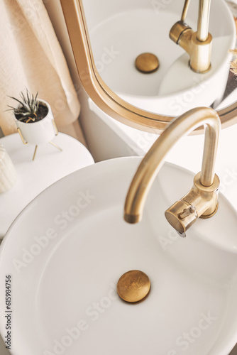 Close-up of an elegant golden faucet in the bathroom sink next to stylish decorations. A beautiful sink with a golden faucet next to an oval mirror and a shelf with hand towels. 