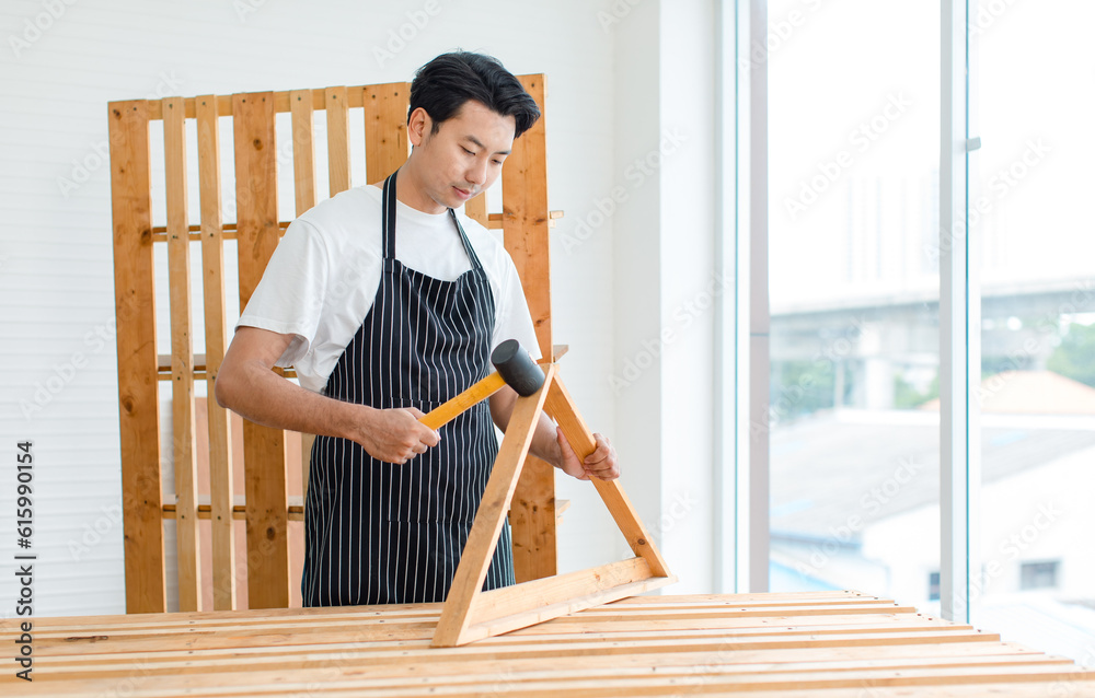 Asian professional skilled male handyman carpenter in apron using rubber hammer pounding on square triangle wood sticks and planks on wooden table workbench in home decoration building workshop Asian professional skilled male handyman carpenter in apron using rubber hammer pounding on square triangle wood sticks and planks on wooden table workbench in home decoration building workshop