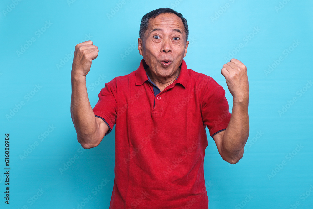 Excited senior man making winner gesture rejoicing with fists isolated over blue background Excited senior man making winner gesture rejoicing with fists isolated over blue background