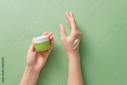 On a green background, hands of female holding a green cream jar unbranded for design and a smear of cream on back of other hand. Scene for advertising cosmetic with minimalist style
