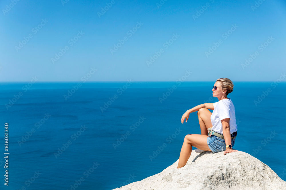 A woman in a white tank top and shorts sits on a high white sandy mountain and looks out over the sea and the landscape below against a bright blue sky. Great view. Tourism, travel and recreation.