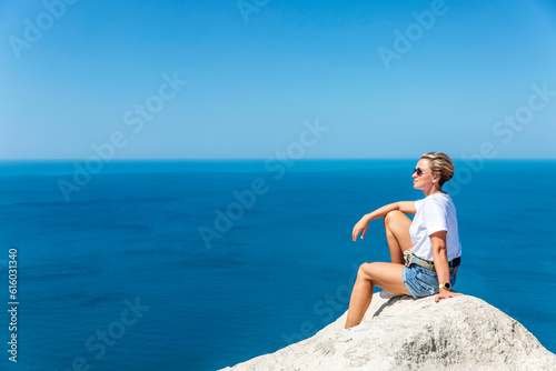 A woman in a white tank top and shorts sits on a high white sandy mountain and looks out over the sea and the landscape below against a bright blue sky. Great view. Tourism, travel and recreation. © Анна Демидова