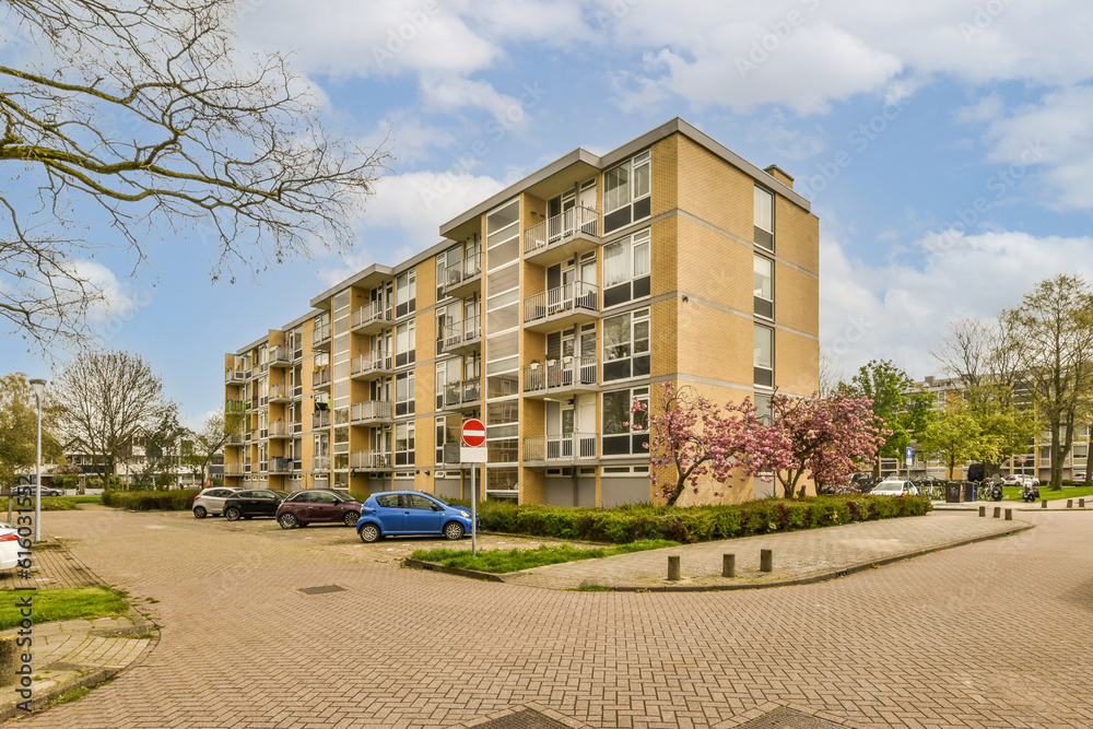 an apartment complex with cars parked on the street and trees in blooming front yard behind it, under a cloudy blue sky an apartment complex with cars parked on the street and trees in blooming front yard behind it, under a cloudy blue sky