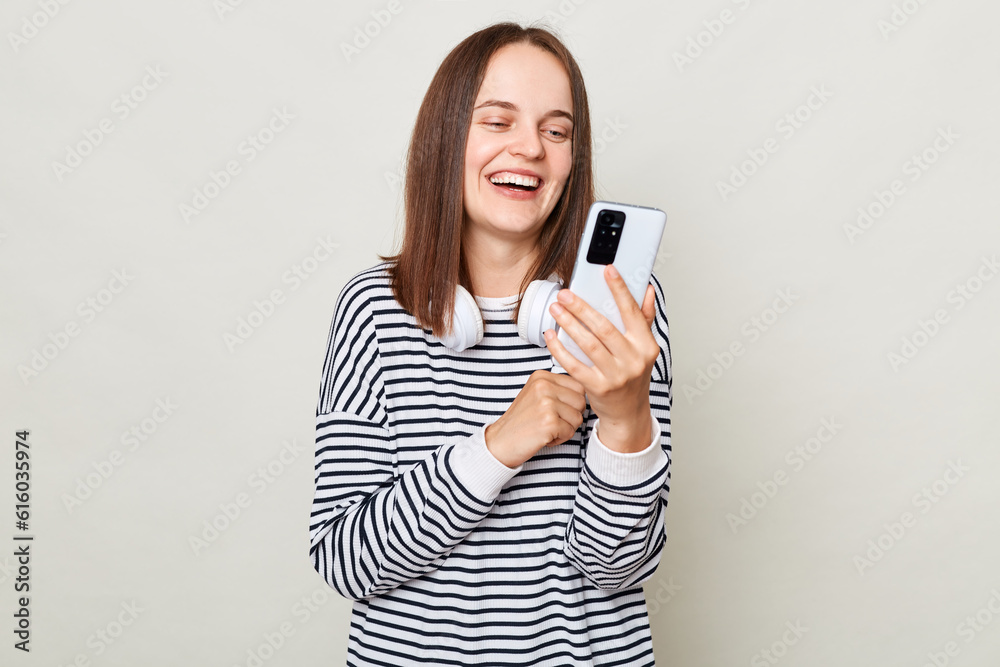 Smiling laughing woman wearing striped shirt standing with headphones over neck isolated over gray background using mobile phone chatting with friends. Smiling laughing woman wearing striped shirt standing with headphones over neck isolated over gray background using mobile phone chatting with friends.