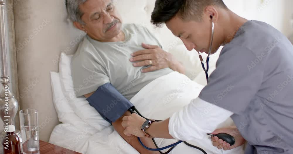 Diverse male doctor testing blood pressure of senior male patient in bed at home, slow motion Diverse male doctor testing blood pressure of senior male patient in bed at home, slow motion