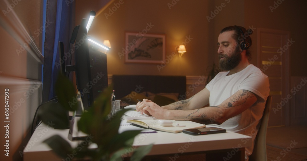 Concentrated financial analyst or trader with pen in hand types keyboard and makes notes. Table illuminated by LED lamps. Working at home concept. Concentrated financial analyst or trader with pen in hand types keyboard and makes notes. Table illuminated by LED lamps. Working at home concept.