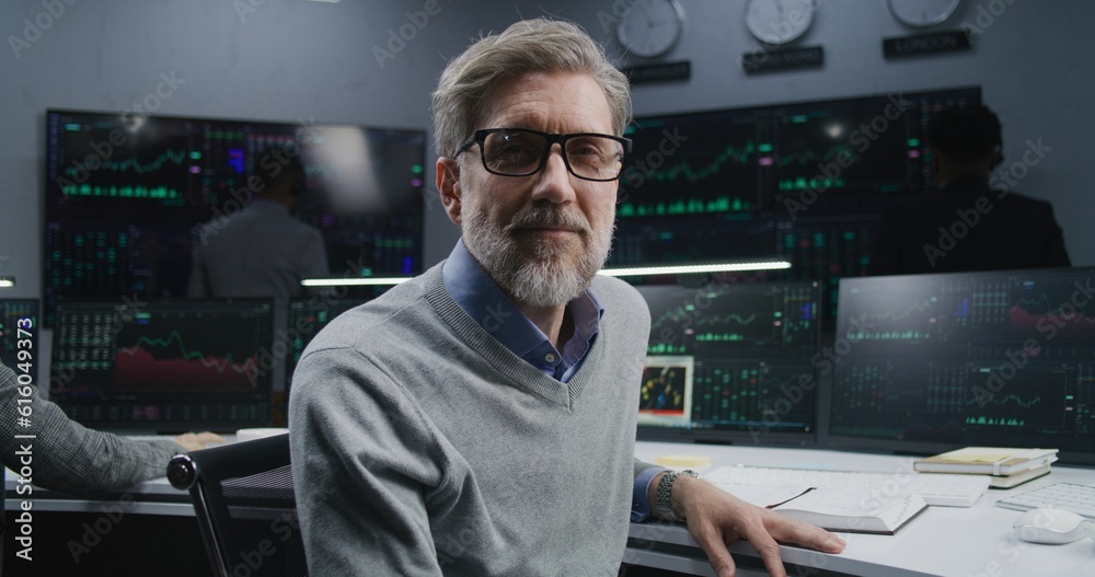 Mature financial analyst works at PC with displayed real-time stocks, turns and looks at camera. Colleagues analyze exchange market charts on big screens at background. Investment concept. Portrait. Mature financial analyst works at PC with displayed real-time stocks, turns and looks at camera. Colleagues analyze exchange market charts on big screens at background. Investment concept. Portrait.