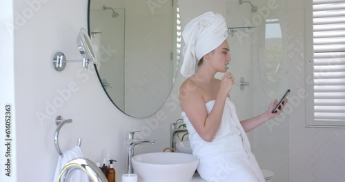 Caucasian woman brushing teeth and using smartphone with towel on head in bathroom in slow motion photo