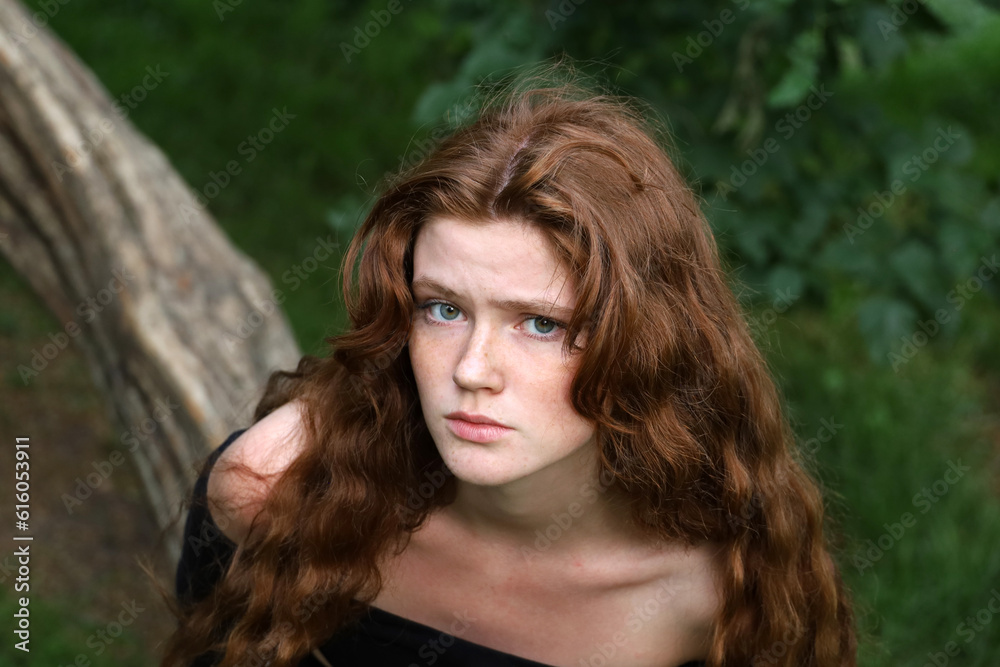 Naklejka premium Portrait of upset girl with long red hair and freckles in summer park. Offended, fright and sad mood