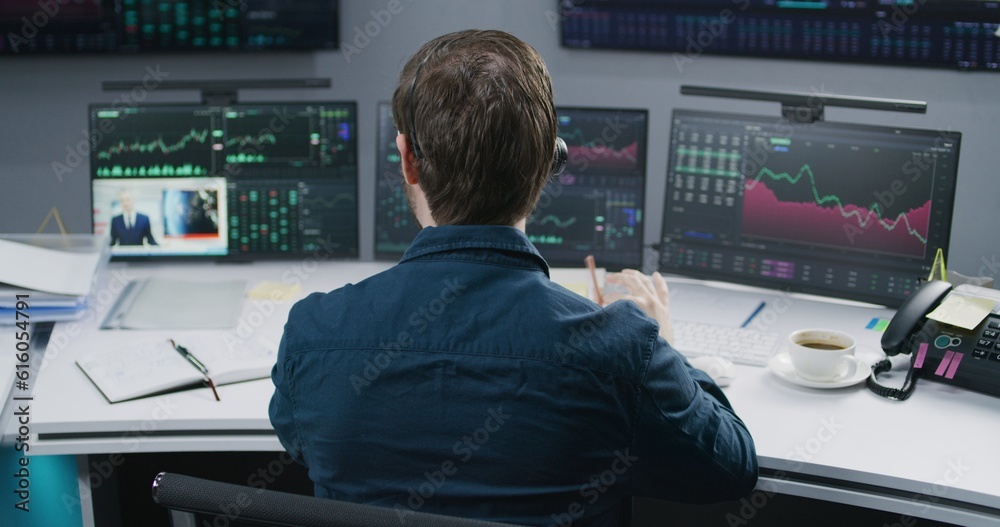 Male analyst in headset watches news, works in bank office on multi-monitor computer with real-time stocks, exchange market charts and commodities. Cryptocurrency trading, investment and analytics.