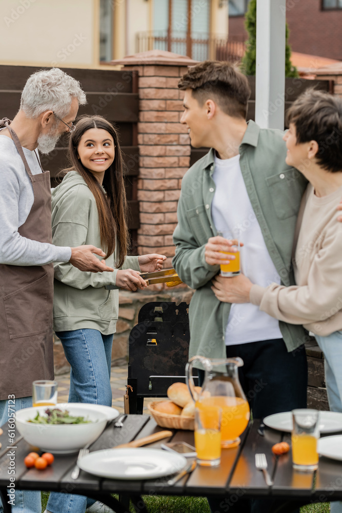 modern parenting, happy parents day, father and teenage daughter preparing food on bbq grill, mother hugging with cheerful young adult son holding glass of orange juice, blurred foreground, backyard modern parenting, happy parents day, father and teenage daughter preparing food on bbq grill, mother hugging with cheerful young adult son holding glass of orange juice, blurred foreground, backyard