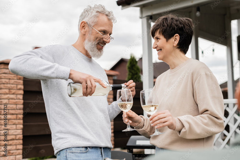 happy marriage, cheerful and bearded middle aged man in glasses pouring white wine into glass of joyful wife, backyard of summer house, romance, casual attire, spending time together happy marriage, cheerful and bearded middle aged man in glasses pouring white wine into glass of joyful wife, backyard of summer house, romance, casual attire, spending time together