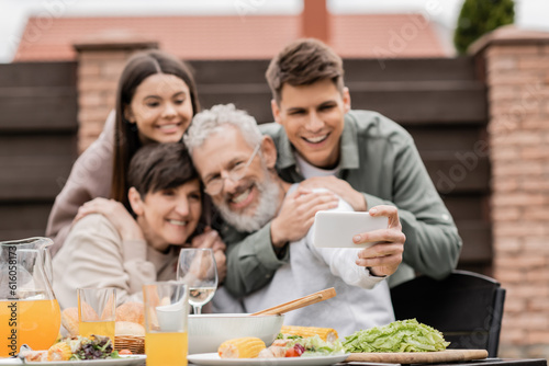 Smartphone in hand of blurred mature man taking selfie with family and kids near summer food during bbq party and parents day celebration at backyard in june  happy parents day concept