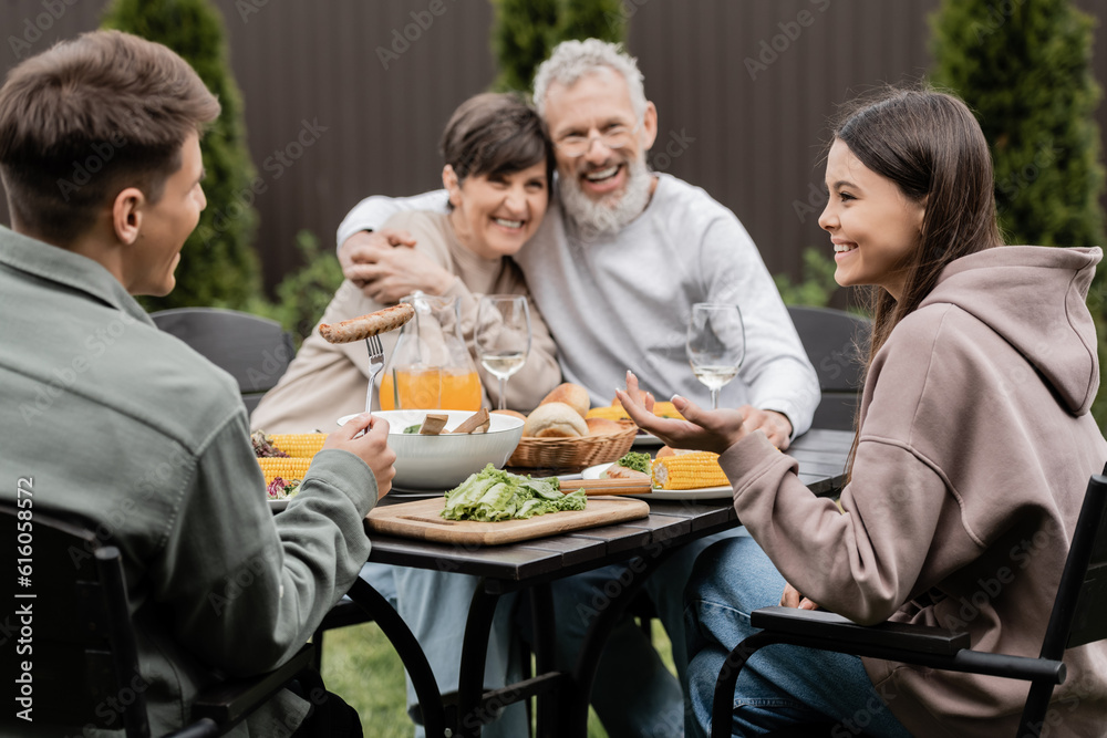 Smiling children talking to each other while sitting near summer food and blurred middle aged parents at background during parents day celebration at background, cherishing family bonds concept