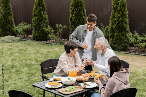 Excited and cheerful middle aged parents toasting with wine glasses near children and summer food during bbq party at backyard  cherishing family bonds concept  spending time together