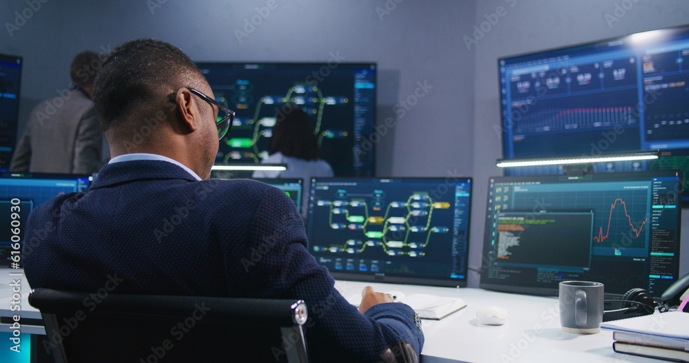 Software programmer sits in front of computer with blockchain network and data server in monitoring office. Team of IT technical specialists work with live analysis charts on big digital screens. Software programmer sits in front of computer with blockchain network and data server in monitoring office. Team of IT technical specialists work with live analysis charts on big digital screens.