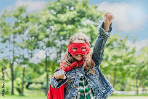 pretty and cute eight-year-old girl wearing a red mask and red cape like a super hero is relaxing and having fun in the park.