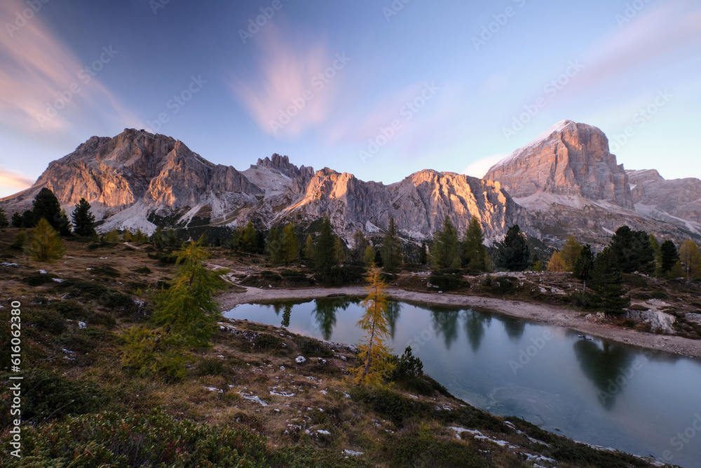 In an alpine setting, the majestic Dolomites are tinged with the warm autumn colors, while the imposing larches are colored with orange and gold, creating reflections on the lake