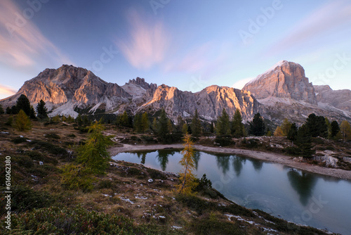 In an alpine setting, the majestic Dolomites are tinged with the warm autumn colors, while the imposing larches are colored with orange and gold, creating reflections on the lake