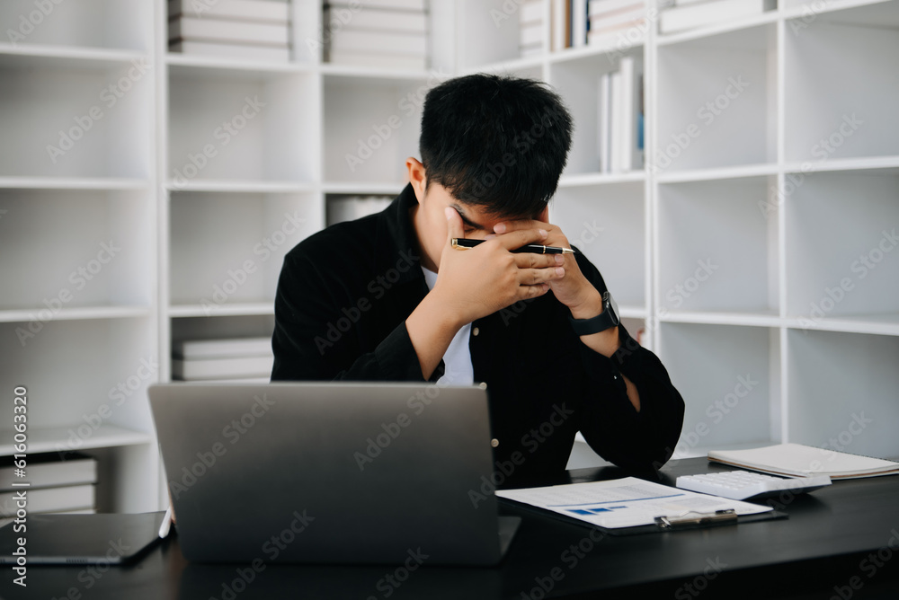 Frustrated young Asian businessman working on a laptop computer sitting at his working place in modern office Frustrated young Asian businessman working on a laptop computer sitting at his working place in modern office