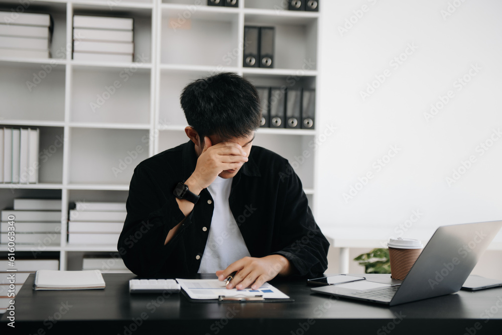 Frustrated young Asian businessman working on a laptop computer sitting at his working place in modern office Frustrated young Asian businessman working on a laptop computer sitting at his working place in modern office