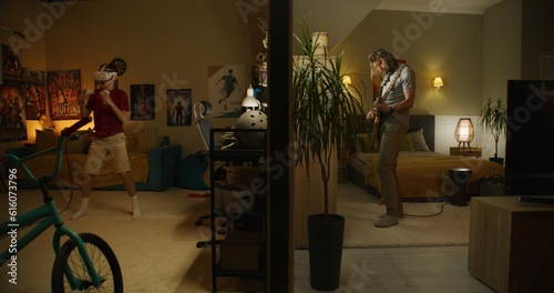 Man plays electric guitar in bedroom. Young boy in VR headset plays augmented reality video game using wireless controllers. View of two rooms separated by wall. Neighbourhood and lifestyle concept.