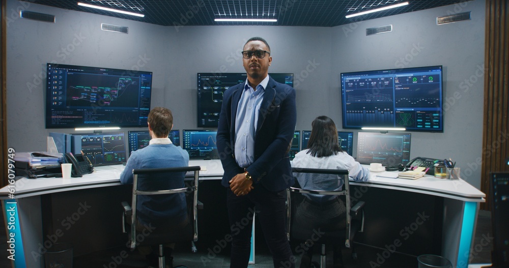 African American IT scientist stands in monitoring room and looks at camera. Multi cultural big data engineers work at computers. Multiple screens with displayed real-time analysis charts. Dolly shot.