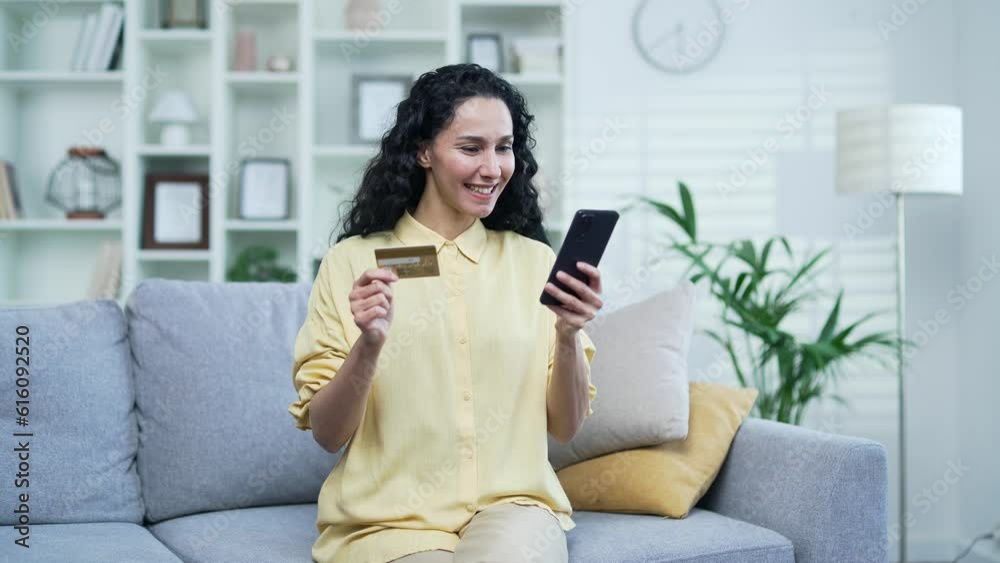 Happy young brunette woman doing online shopping typing credit card number on smartphone while sitting on sofa at home. A smiling female happily makes a purchase, satisfied with the product promotion Happy young brunette woman doing online shopping typing credit card number on smartphone while sitting on sofa at home. A smiling female happily makes a purchase, satisfied with the product promotion