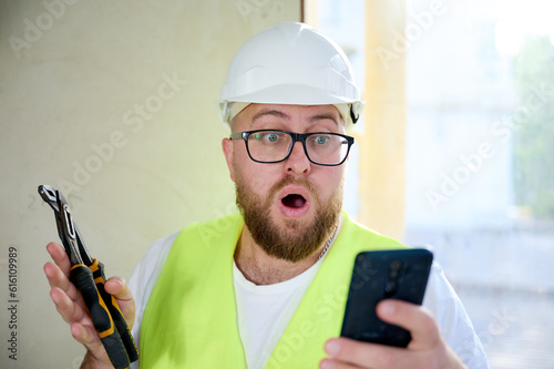 Engineer with white helmet using his smart phone at the construction site. Portrait of construction worker in uniform playing games or makes bets on mobile phone during break. Fast delivery. Win.