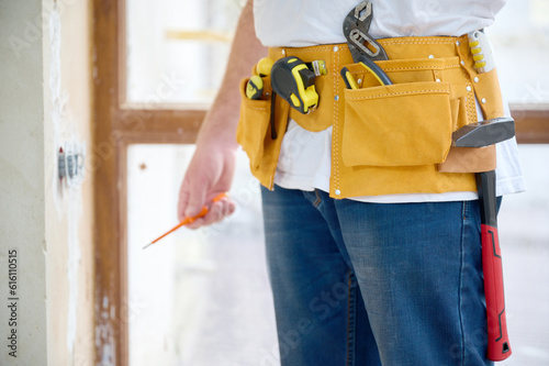 Cropped view of the electrician with a screwdriver in his hand wearing in white T-shirt and blue jeans with yellow tool belt near concrete or cement wall and wide light window.