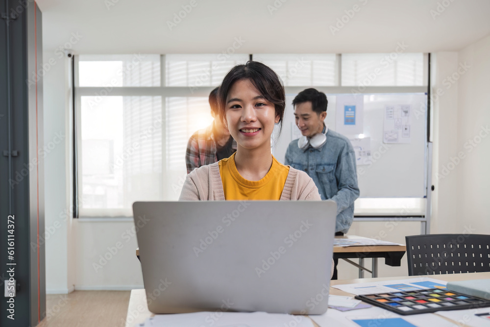 A focused young Asian female marketing assistant is working on her work on her laptop at her desk while her coworkers are working in the room. A focused young Asian female marketing assistant is working on her work on her laptop at her desk while her coworkers are working in the room.