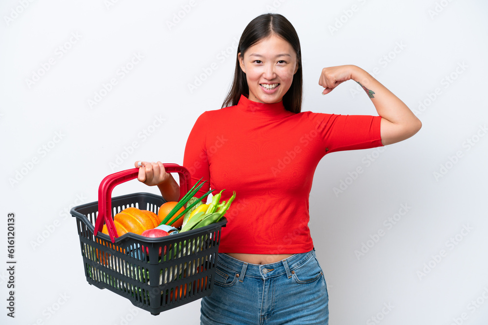 Young Asian woman holding a shopping basket full of food isolated on white background doing strong gesture Young Asian woman holding a shopping basket full of food isolated on white background doing strong gesture