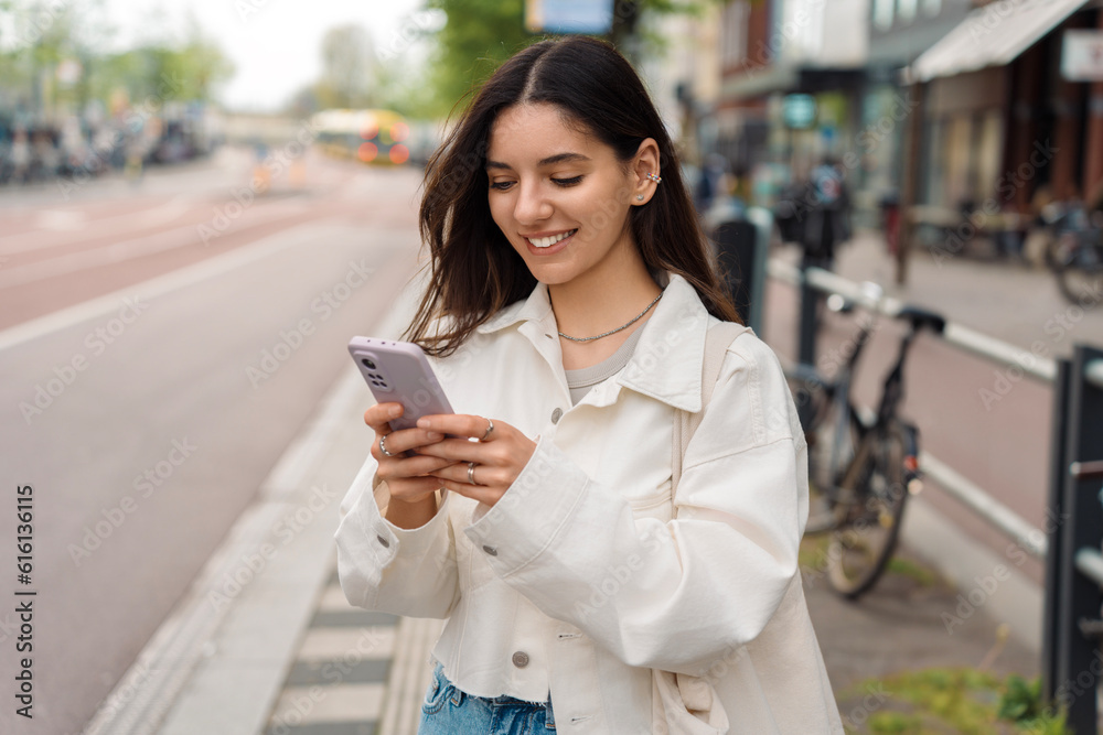 Everyday commute. Cheerful young woman student using the app on her phone while waiting for the public transport