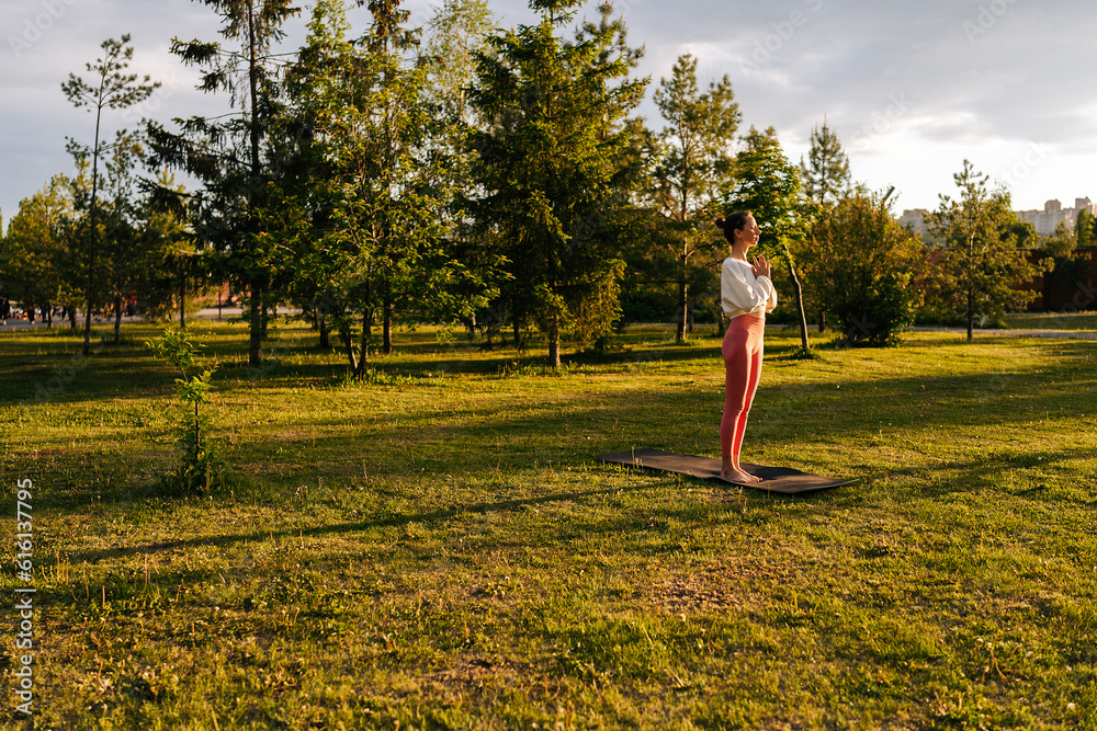 Wide shot of relaxed yogini female with closed eyes practicing yoga performing namaste pose, background of sunbeam green grass on summer park. Female practicing yoga and meditation in morning at dawn. Wide shot of relaxed yogini female with closed eyes practicing yoga performing namaste pose, background of sunbeam green grass on summer park. Female practicing yoga and meditation in morning at dawn.