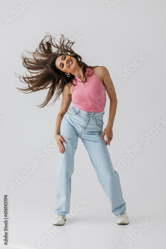 A sporty cute girl in a tank top and jeans dances with pleasure and smiles. Indoor portrait of stunning female model with vintage hairstyle waving her hands against light background.