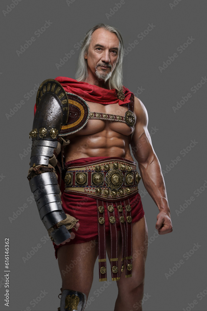 Aged but still mighty, this muscular gladiator with long grey hair and beard stands proudly in lightweight armor against a grey background Aged but still mighty, this muscular gladiator with long grey hair and beard stands proudly in lightweight armor against a grey background