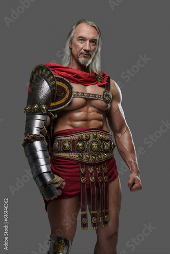 Aged but still mighty, this muscular gladiator with long grey hair and beard stands proudly in lightweight armor against a grey background