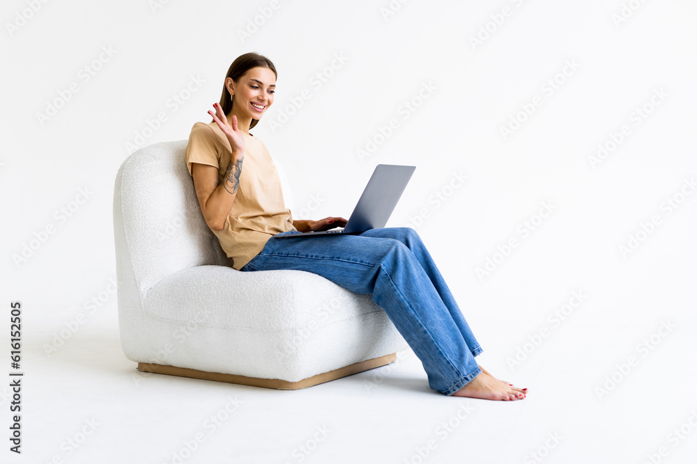 Cheery woman in headphones having online video call on laptop, sitting on bag chair against white background Cheery woman in headphones having online video call on laptop, sitting on bag chair against white background