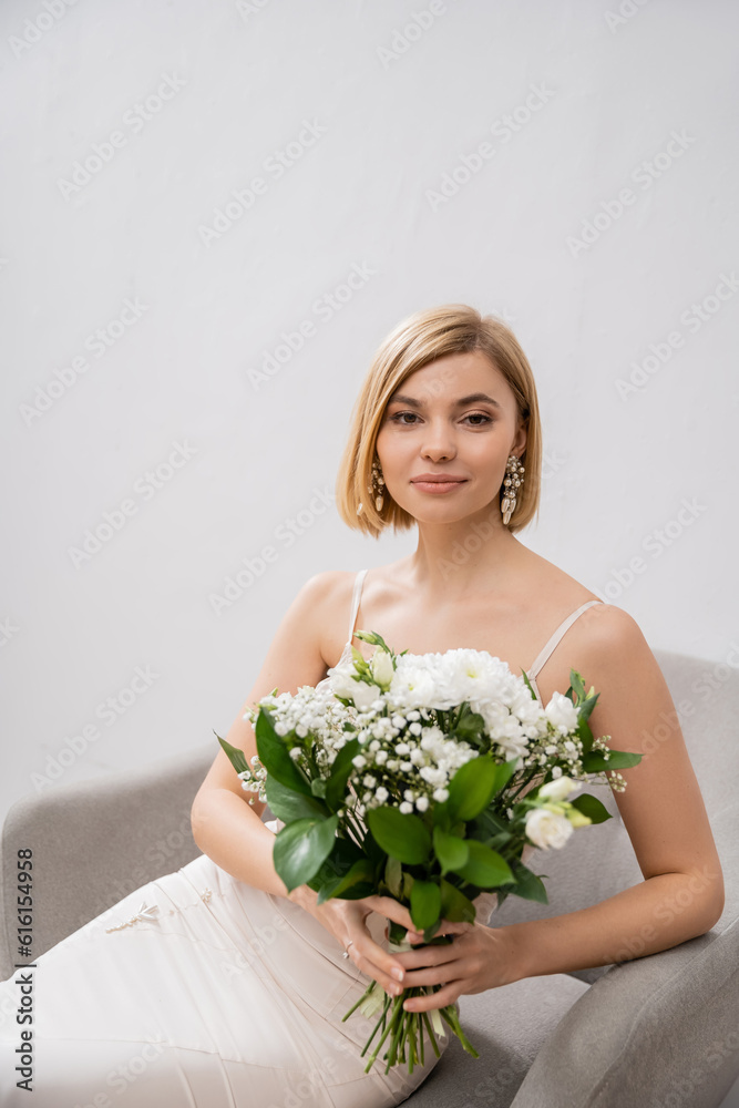elegant and blonde bride in wedding dress sitting in armchair and holding bouquet on grey background, white flowers, bridal accessories, happiness, special occasion, beautiful, feminine, blissful elegant and blonde bride in wedding dress sitting in armchair and holding bouquet on grey background, white flowers, bridal accessories, happiness, special occasion, beautiful, feminine, blissful