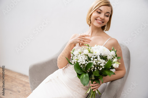 cheerful and elegant bride in wedding dress sitting in armchair and holding bouquet on grey background, white flowers, bridal accessories, happiness, special occasion, beautiful, feminine, blissful