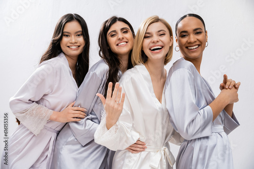 bridal shower  four women  happy bride showing engagement ring near interracial bridesmaids in silk robes  cultural diversity  having fun together  friendship goals  brunette and blonde women