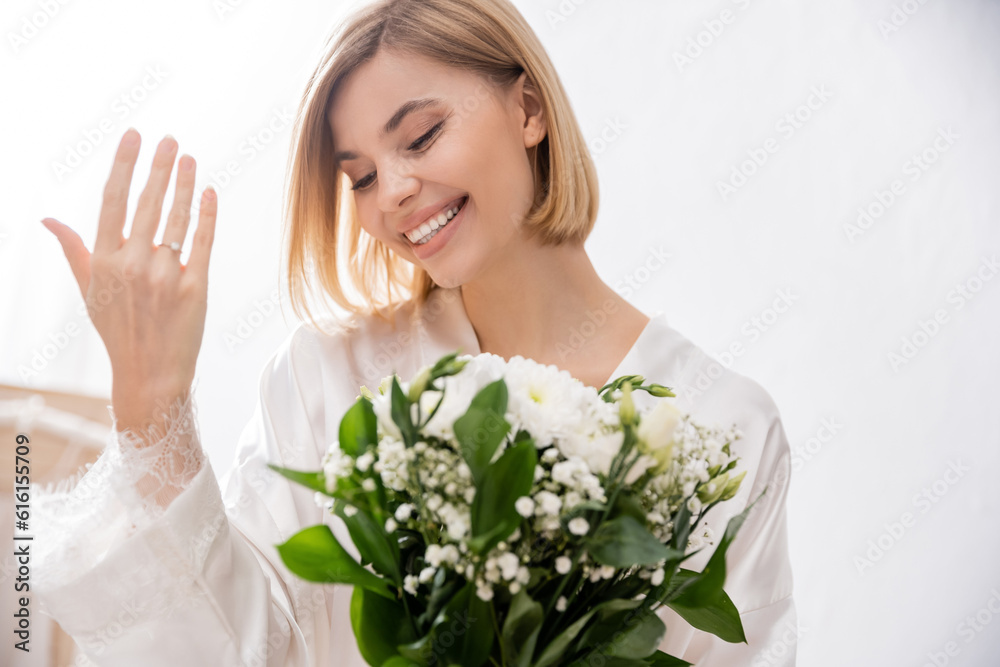 happiness, cheerful bride with blonde hair wearing white silk robe and holding bridal bouquet, showing engagement ring, young woman, beautiful, excitement, feminine, blissful, white flowers