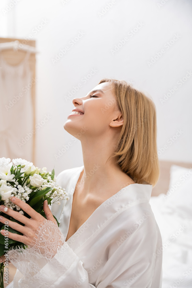 happiness, cheerful bride with blonde hair sitting on bed and holding bridal bouquet, young woman in white robe, beautiful, excitement, feminine, blissful, wedding preparation, white flowers happiness, cheerful bride with blonde hair sitting on bed and holding bridal bouquet, young woman in white robe, beautiful, excitement, feminine, blissful, wedding preparation, white flowers