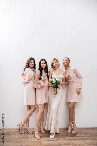 positivity, blonde bride in wedding dress holding bouquet and standing near interracial bridesmaids with champagne glasses on grey background, racial diversity, fashion, multicultural young women