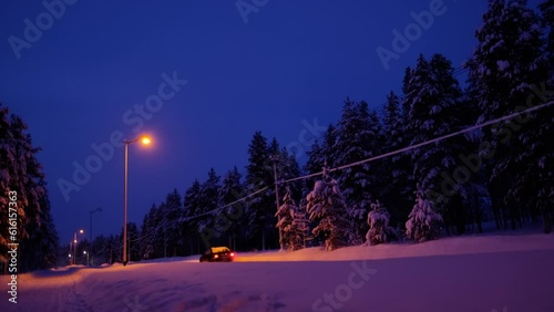 evening snowy road, car is passing by, shining headlights, thick layer of snow on trees, autotourism in Lapland, polar night, snowfall in forest, seasonal change of weather concept, beauty of Arctic photo