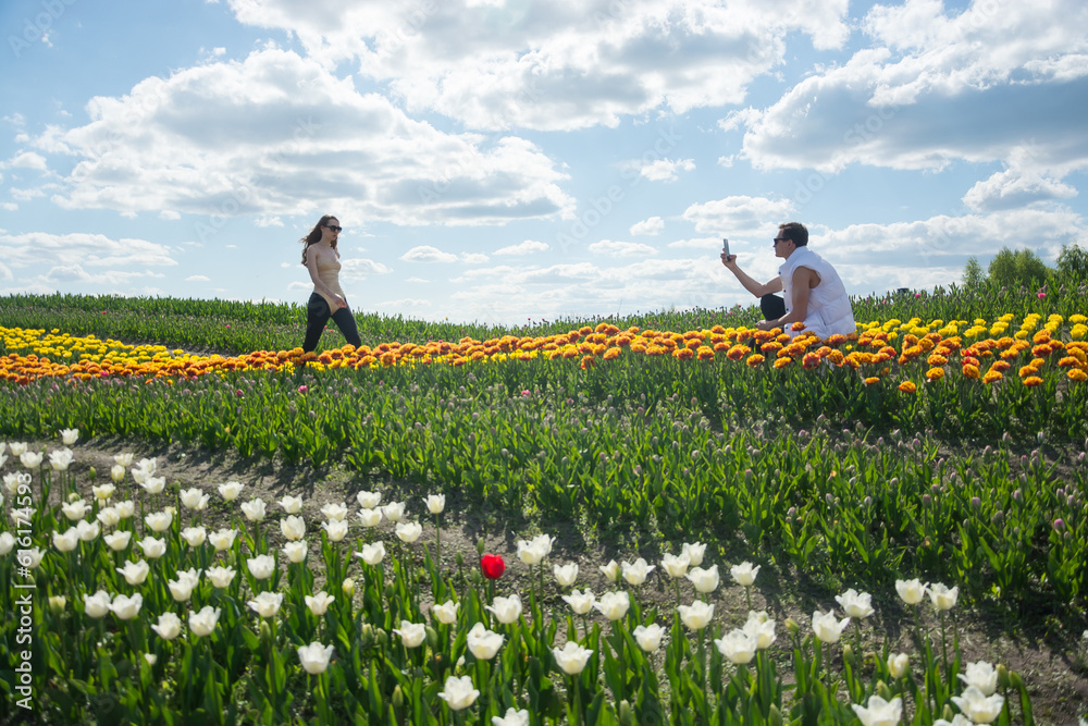 A guy takes a picture of his girlfriend with his phone at field of colorful tulips background.