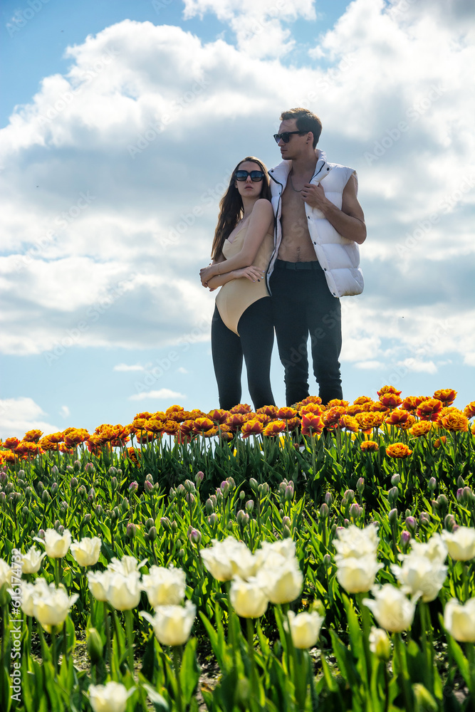 A beautiful young couple, fashion models stands in sportswear at the background of a field of colorful tulips.